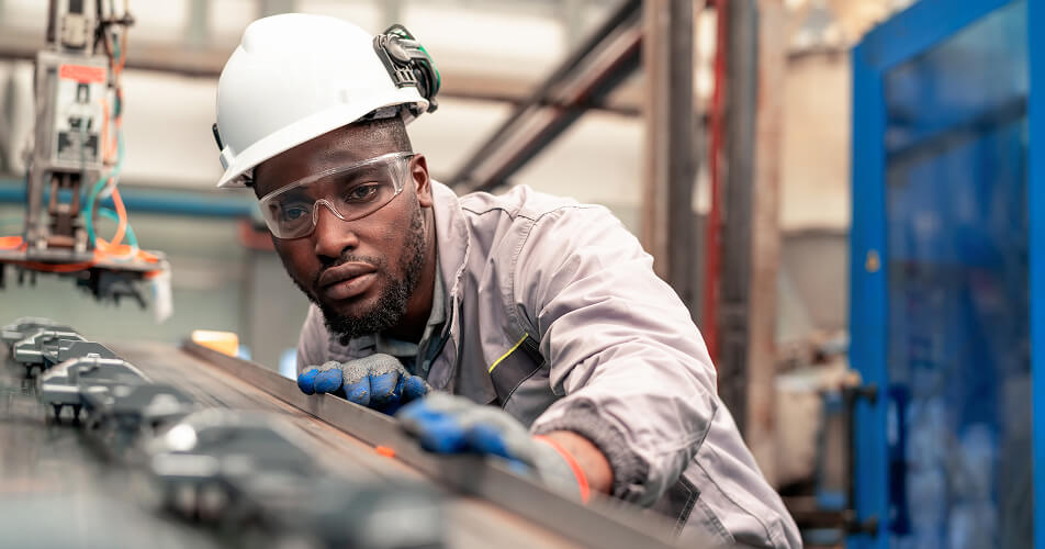 An engineer in a factory wearing protective equipment leans close to the production line to check the quality of the components.