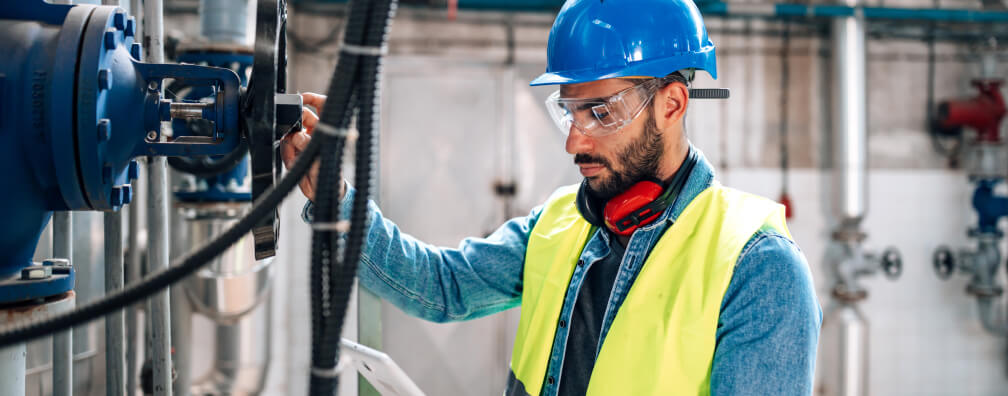 An engineer wears a blue hard hat, safety goggles and a high-visibility vest while inspecting industrial pipes and machinery in a large mechanical room.
