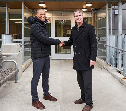 Two professionals shaking hands and smiling in front of an apartment building entrance.