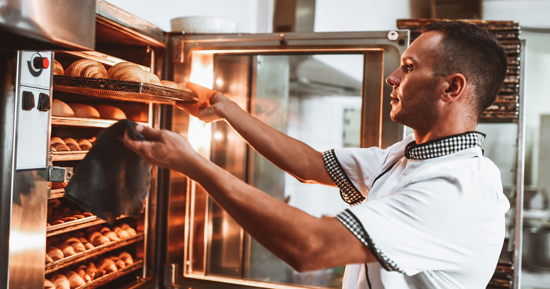 A cook, viewed from the side, removes a shallow sheet pan of baked goods from an ENERGY STAR® certified rack oven.