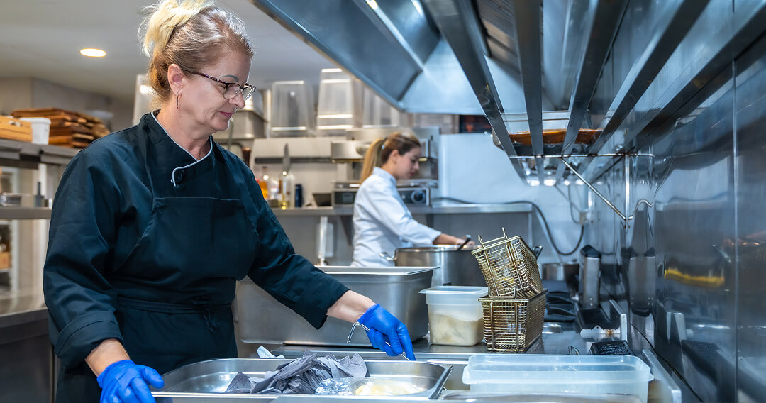 Two cooks prepare meals in a commercial kitchen. The woman in the foreground wears a black chef’s jacket, blue gloves, and glasses while preparing food at a stainless steel counter. In the background, the other cook-this one wearing white chef’s coat-works at a different station. Modern stainless steel appliances, containers, and other kitchen equipment are visible around them.