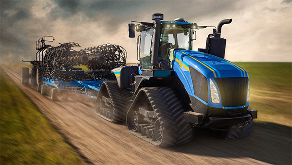 A large blue tractor pulls a piece of seeding equipment down a dirt road, kicking up dust as it moves through an open rural landscape under a cloudy sky.
